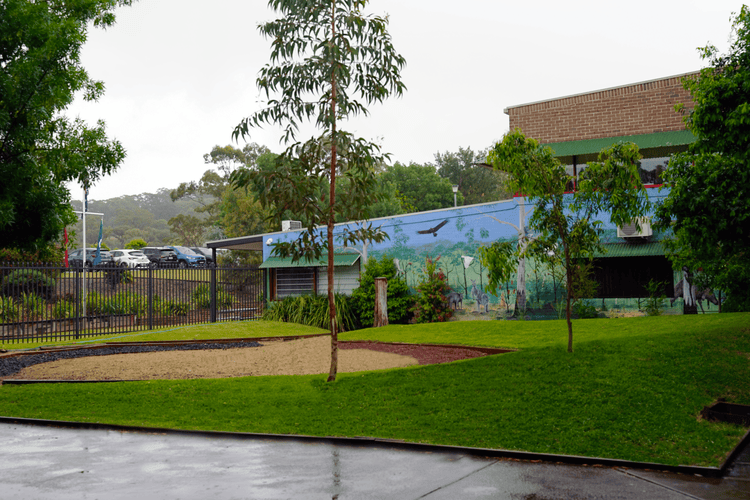 The Yarning Circle featuring a mural in the background, three circular areas made of different natural materials, surrounded by grass and two trees.