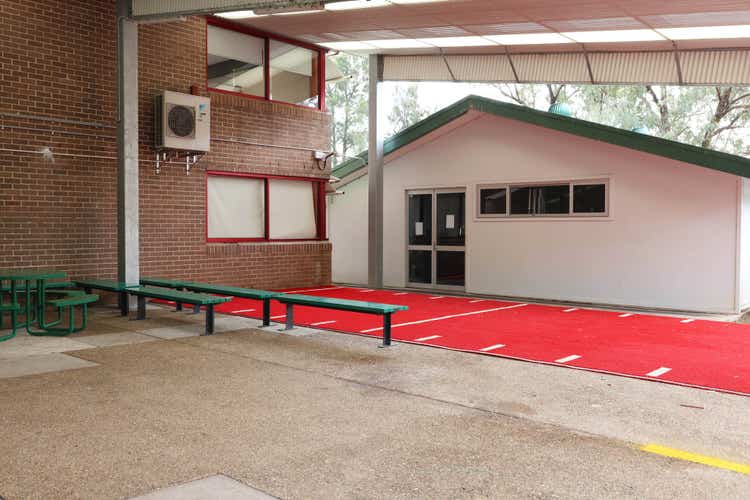 The Gym COLA showing the side of a brick building next to a white building, with synthetic red turf and concrete in front, featuring green bench seating and a green table with chairs.