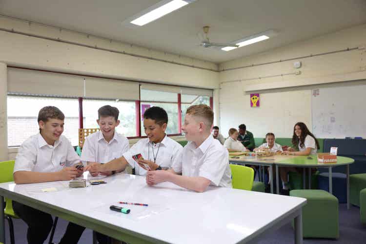 A group of boys in school uniforms sitting at a table playing cards, while a group of girls play Jenga in the background.