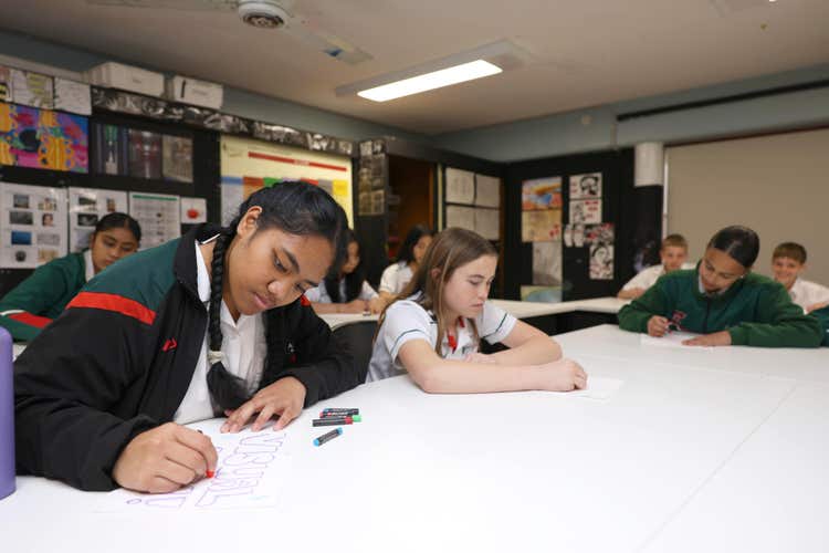 Students drawing with crayons at tables in an art room.
