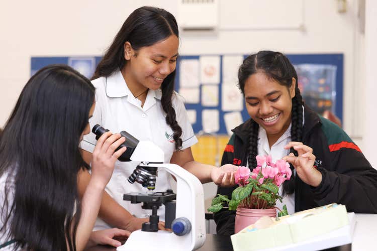 Three girls in a science lab, two examining potted flowers and the third looking through a microscope.