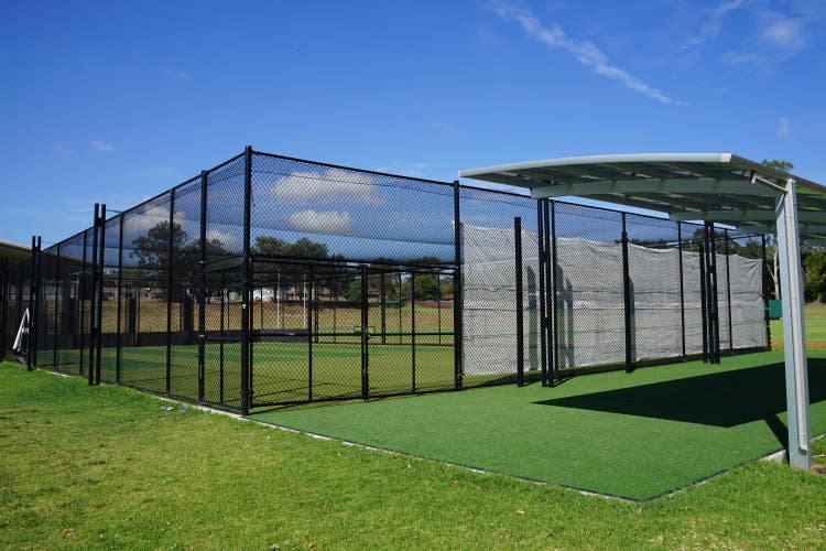 An enclosed cricket pitch with three synthetic grass lanes and a covered viewing area outside the enclosure.