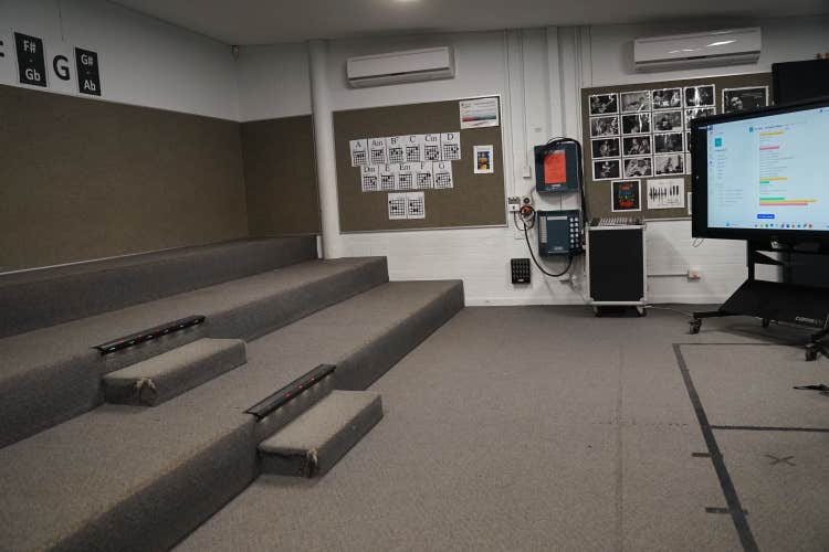 A room with tiered carpeted seating, a TV screen display, music equipment along the wall, and various music posters on the walls.