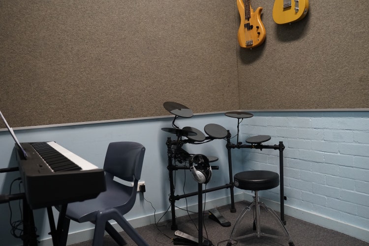 A rehearsal room with a keyboard, a drum set with a stool, two guitars hanging on the wall, headphones on a stand, and a seat at the keyboard.